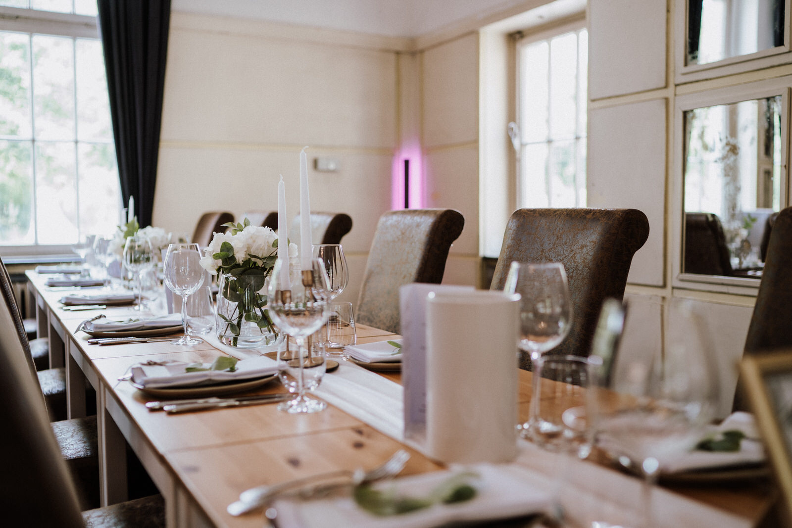 ang gedeckter Tisch in einem hellen Veranstaltungsraum, mit Holztisch, gepolsterten Stühlen, Gläsern, Kerzen und schlichter weißer Blumendekoration.