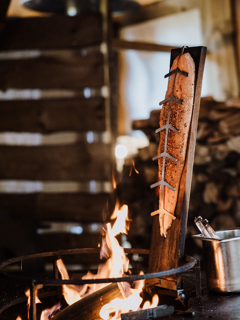Lachsfilet auf Holzplanke über offenem Feuer, rustikale Zubereitung mit Flammen im Vordergrund