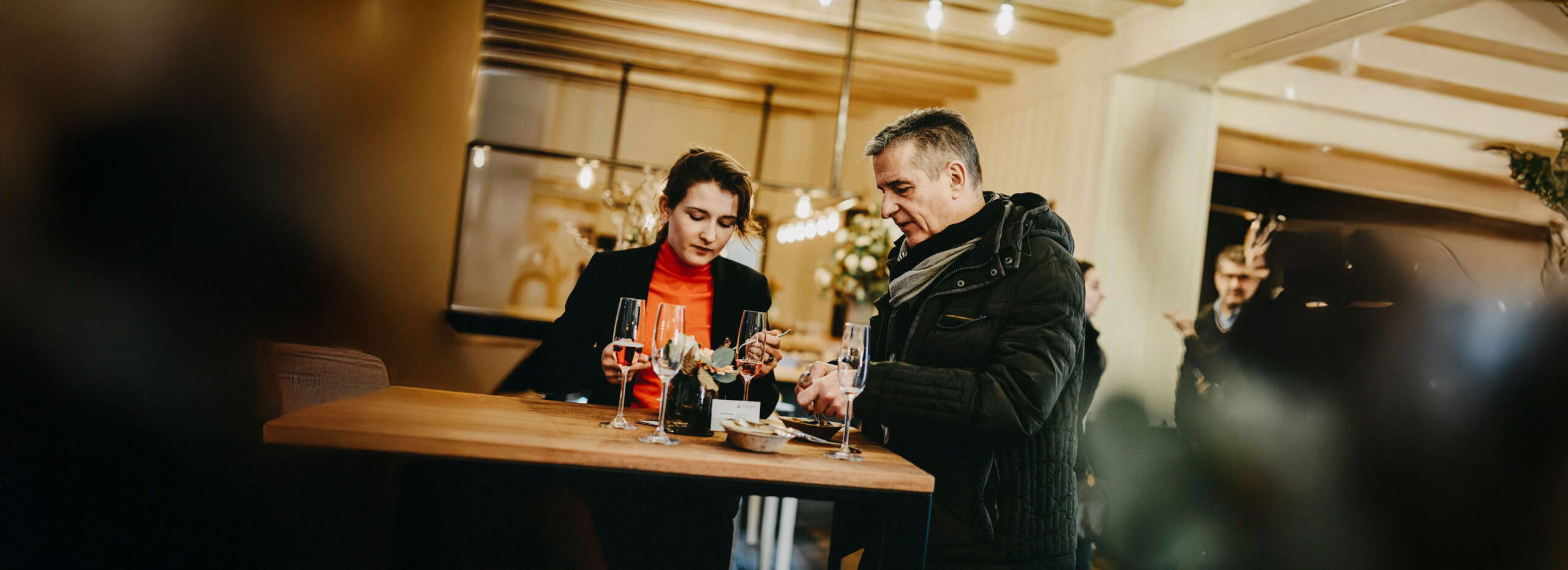 Zwei Personen stehen an einem Holztisch, trinken Sekt und unterhalten sich bei einem Firmenevent in stilvollem Innenraum.