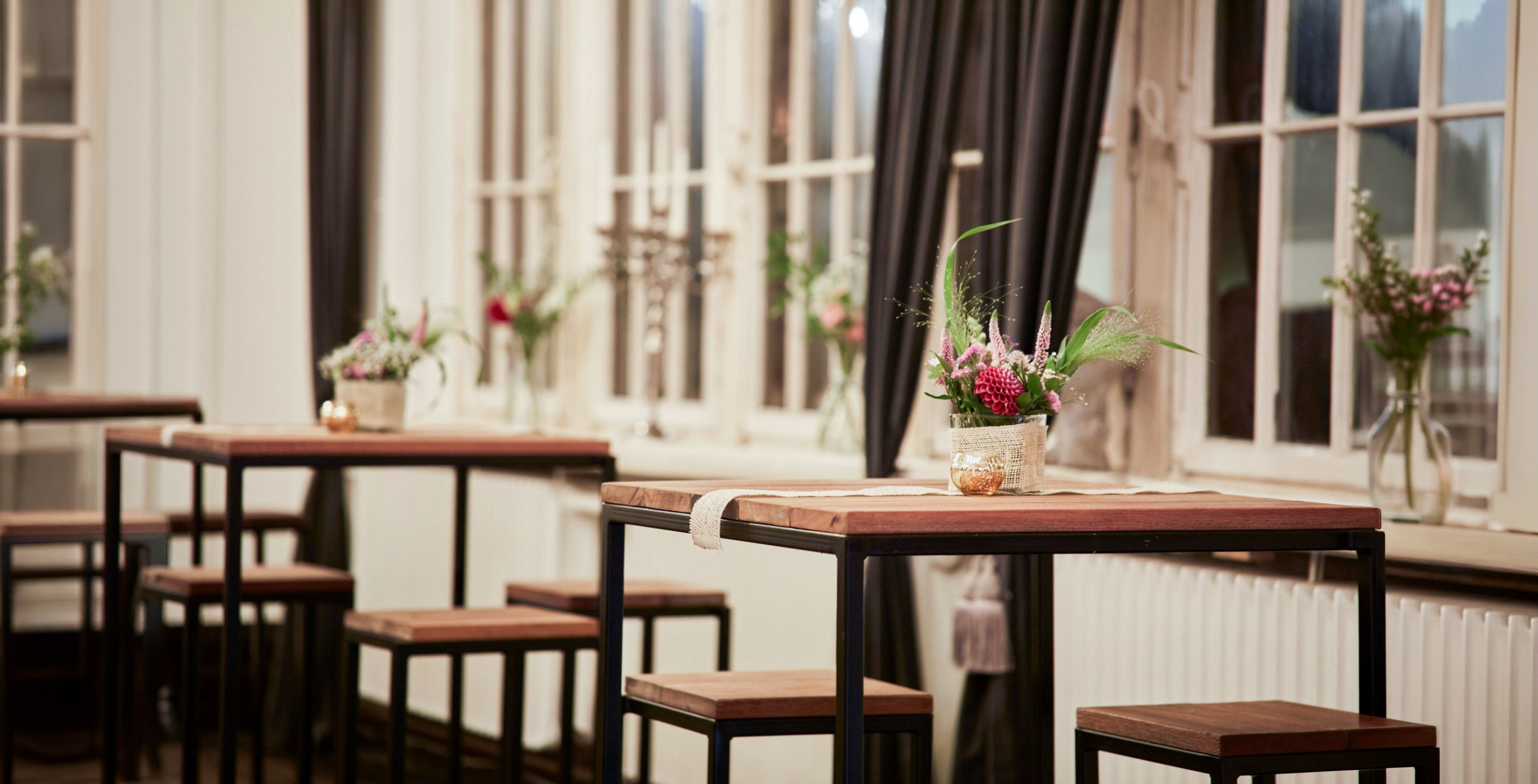 Stehtische mit Blumenvasen vor großen Fenstern in elegantem Veranstaltungsraum