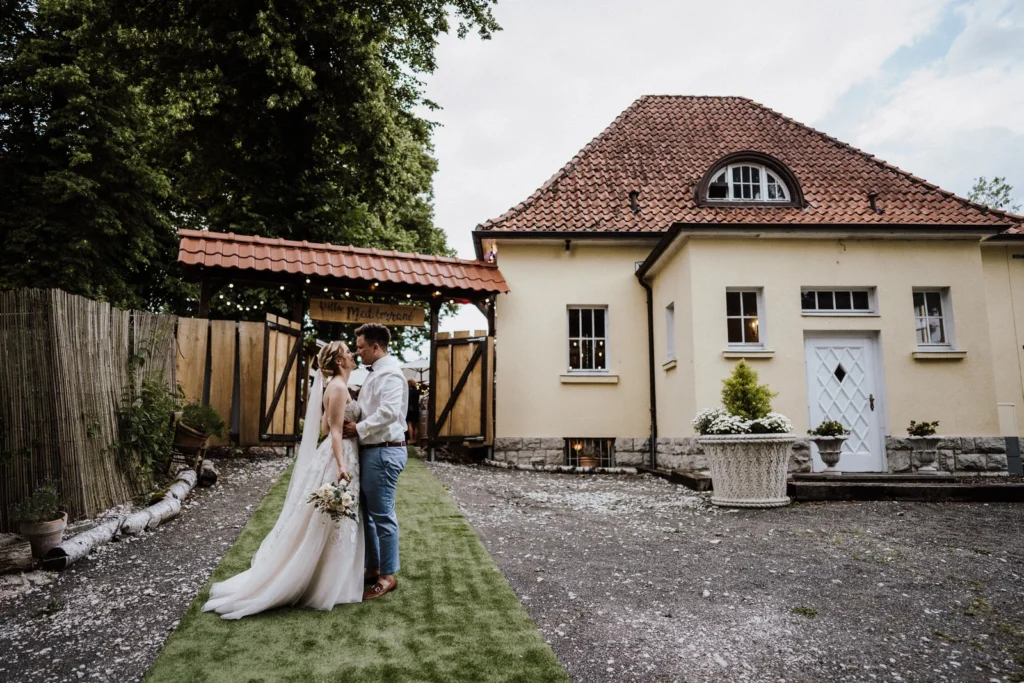 Brautpaar steht sich verliebt gegenüber im Außenbereich vor der Villa, mit grünem Weg, Holztor und dem Gebäude der Location im Hintergrund.