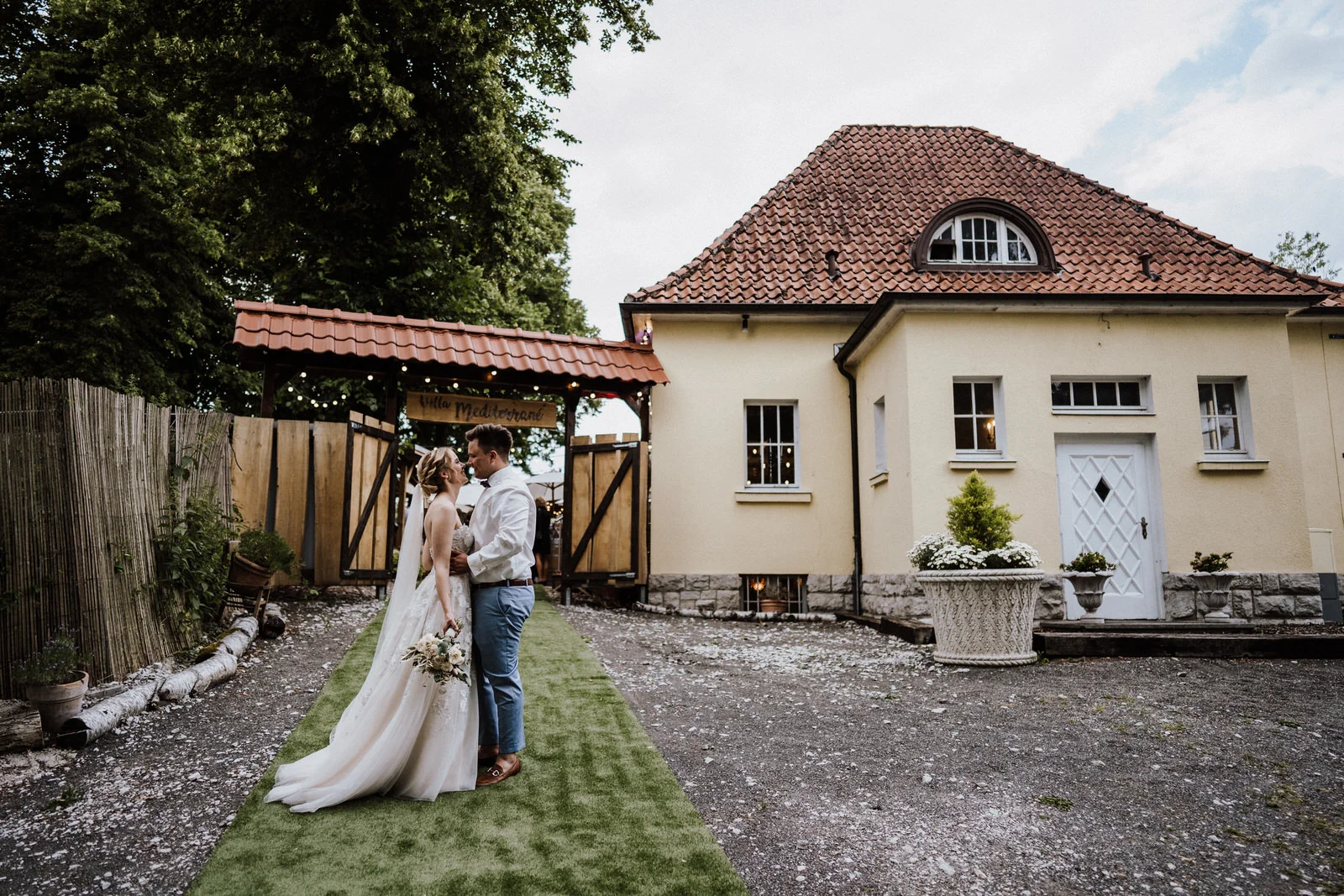 Brautpaar steht sich verliebt gegenüber im Außenbereich vor der Villa, mit grünem Weg, Holztor und dem Gebäude der Location im Hintergrund.