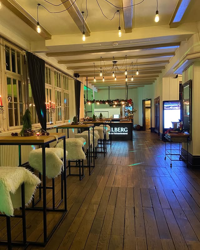 Stimmungsvoll beleuchteter Innenraum einer Eventlocation mit hohen Stehtischen, Bar im Hintergrund und warmem Licht“